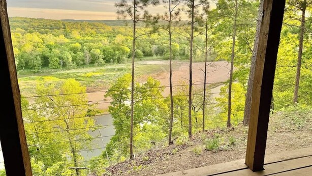 Blick durch ein Fenster mit Holzrahmen auf eine Landschaft mit Bäumen, einem Fluss und hügeligem Gelände.