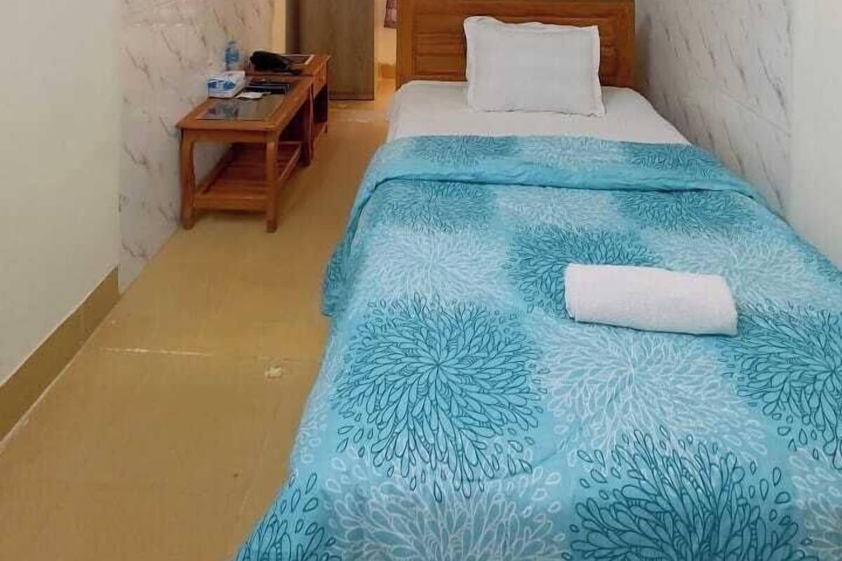A small, well-lit room with a bed, a wooden nightstand, and a desk.