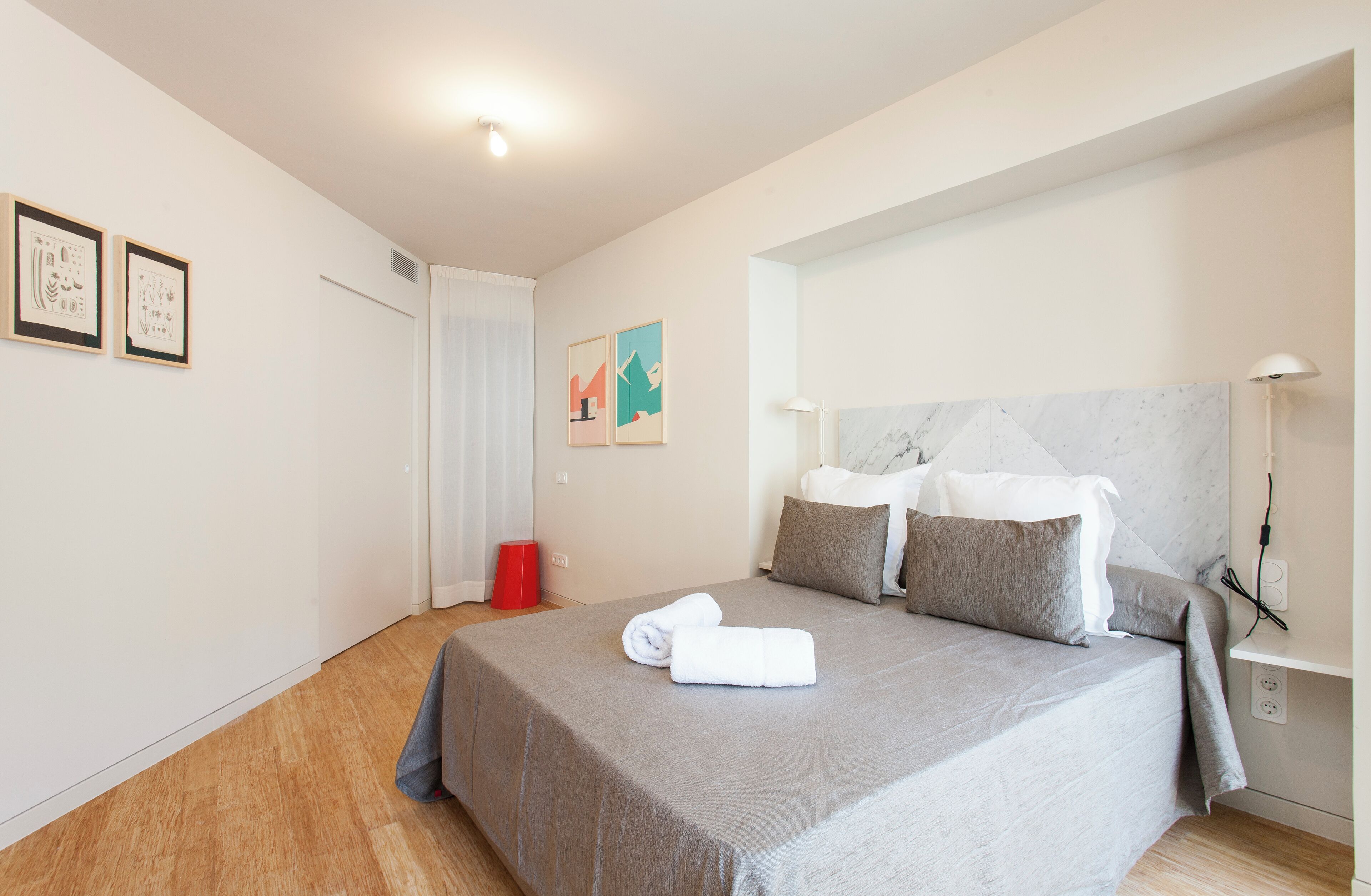A neatly made bed with a grey bedspread and pillows, a red trash bin, and a wall with framed artwork.