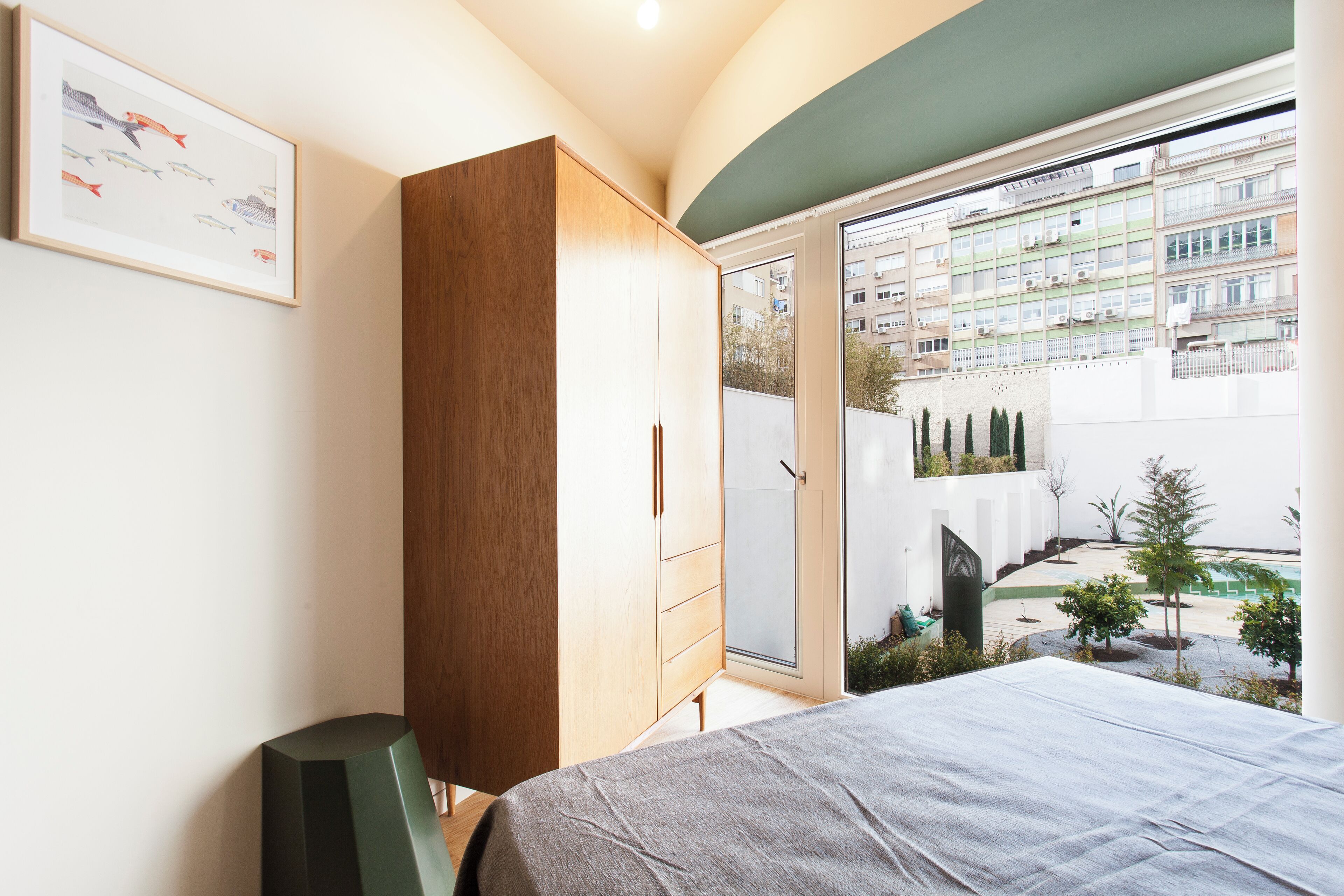 A bedroom with a bed, wooden wardrobe, and a large window with a view of a residential area.