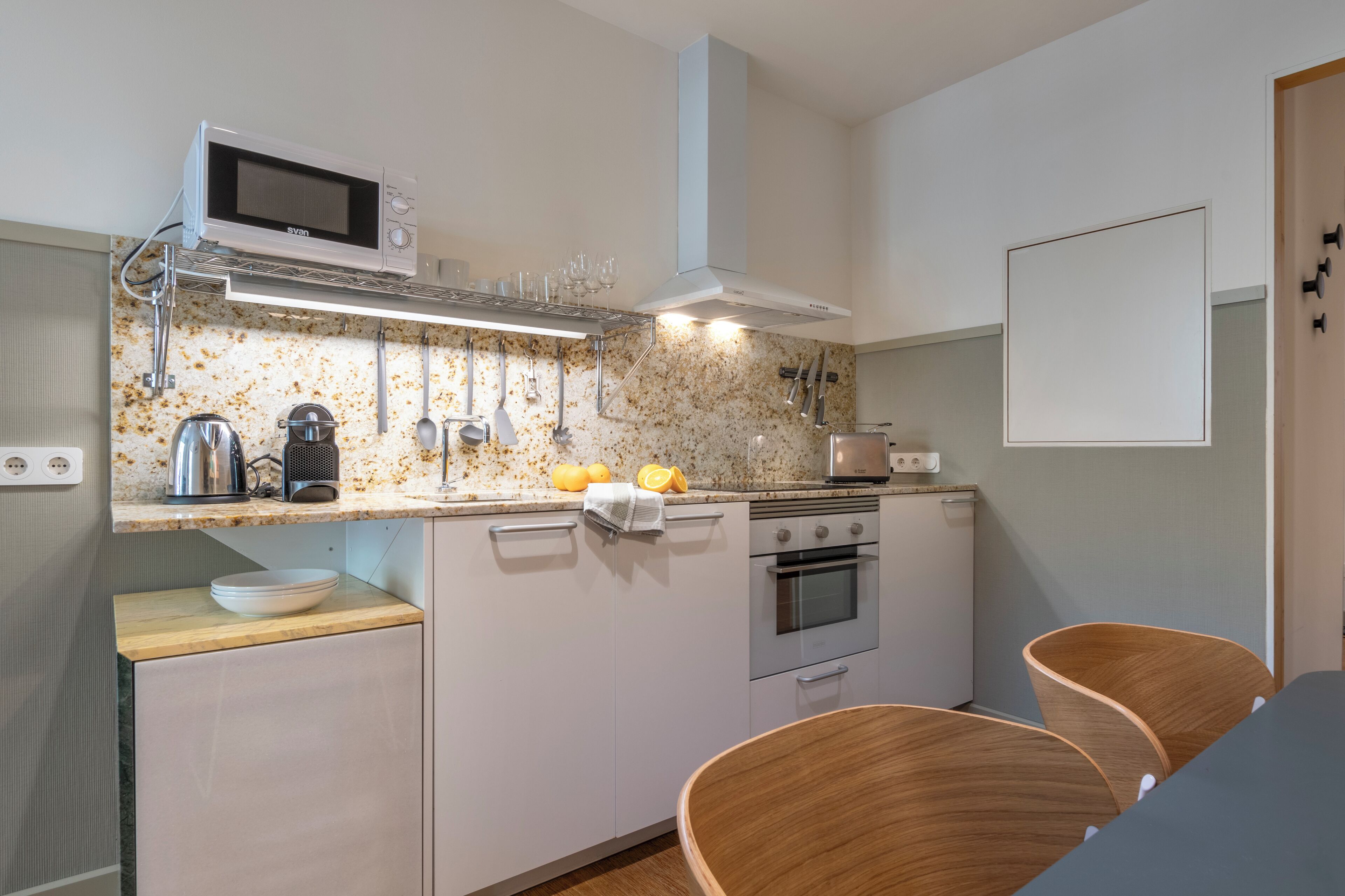 A compact kitchen with a microwave, kettle, and toaster. There are two wooden chairs and a small table.