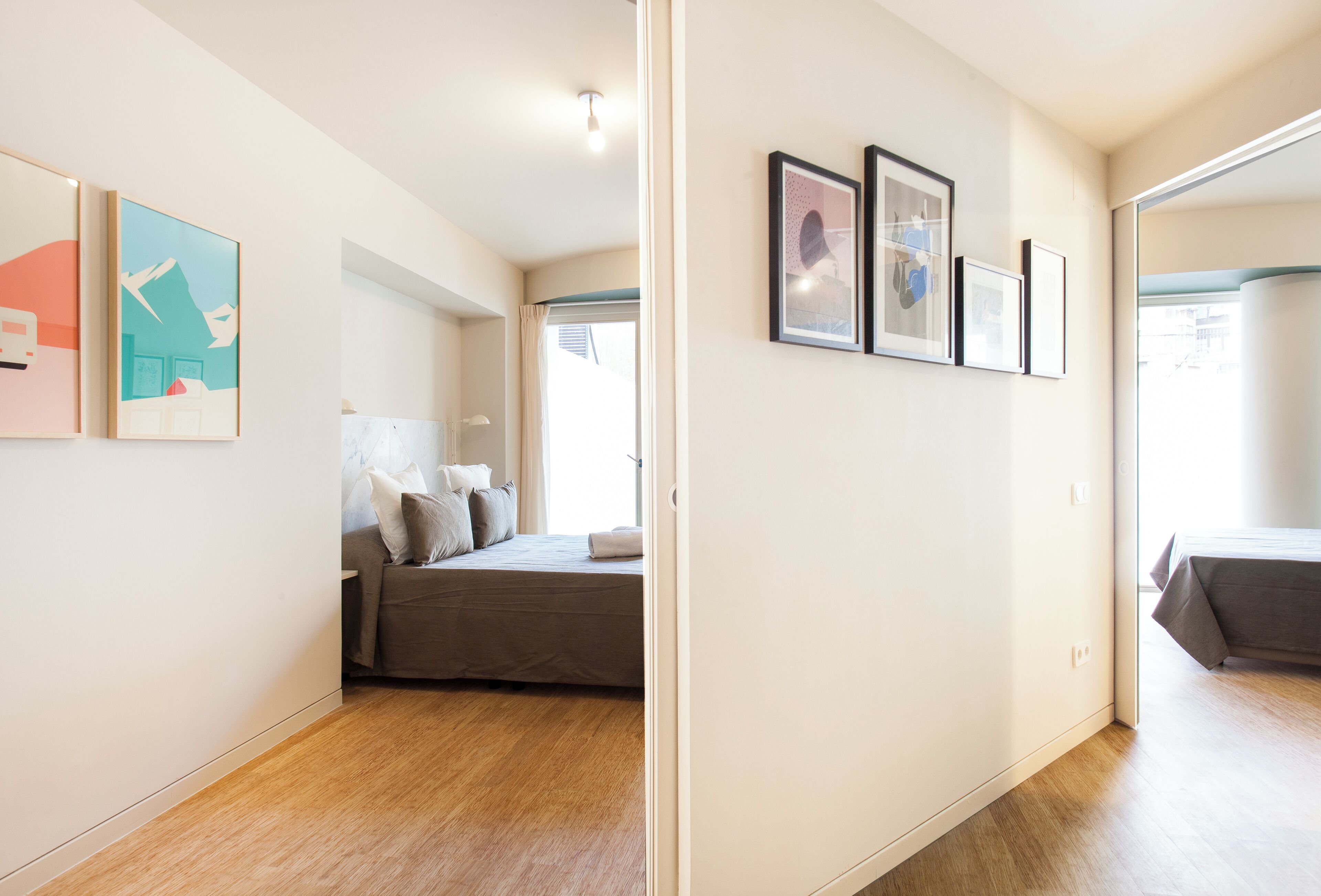 A modern bedroom with a bed, a sofa, and a large window. There are framed pictures on the wall and a door leading to another room.