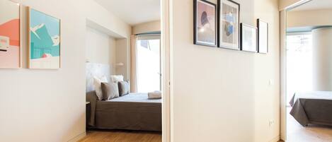 A modern bedroom with a bed, a sofa, and a large window. There are framed pictures on the wall and a door leading to another room.