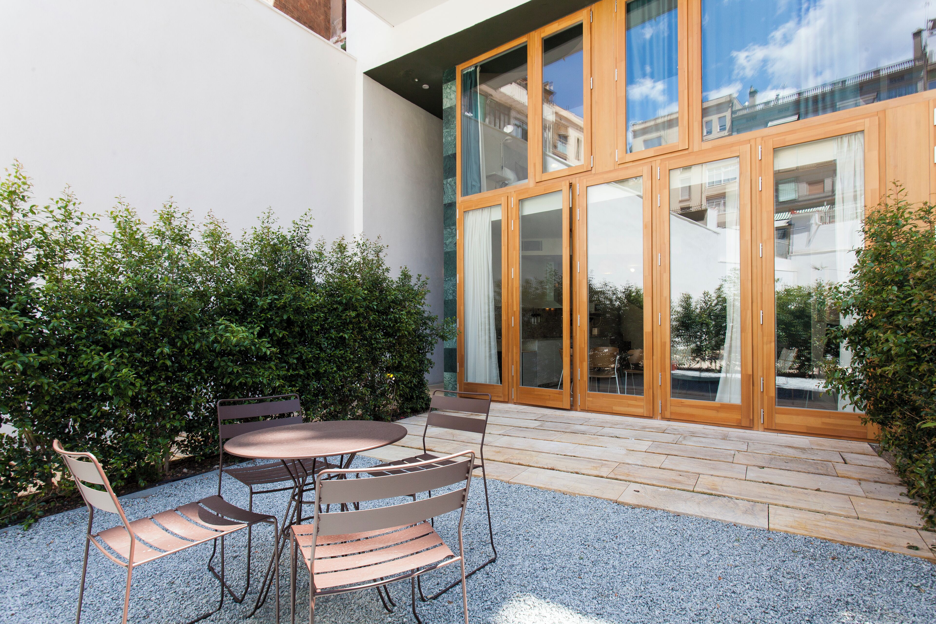 A patio with a table and chairs, surrounded by greenery and a building with large glass doors.