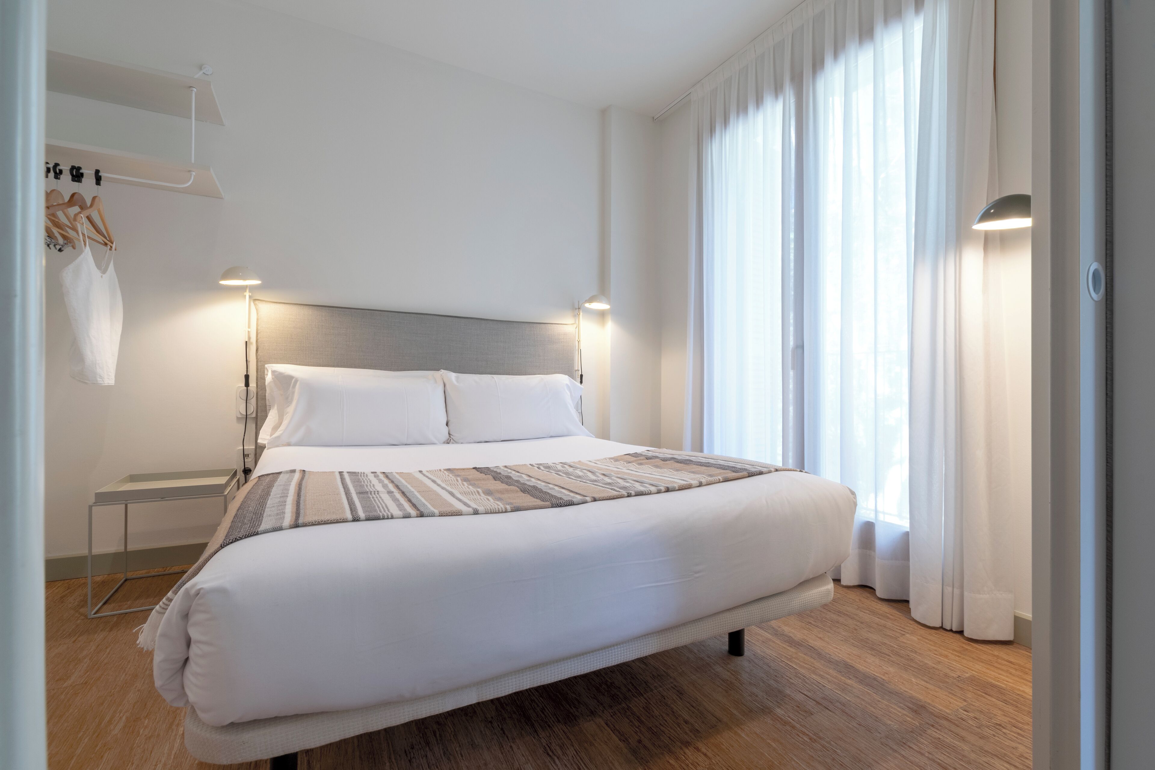 A modern hotel room with a large bed, bedside tables, and a view of the outdoors through sheer curtains.