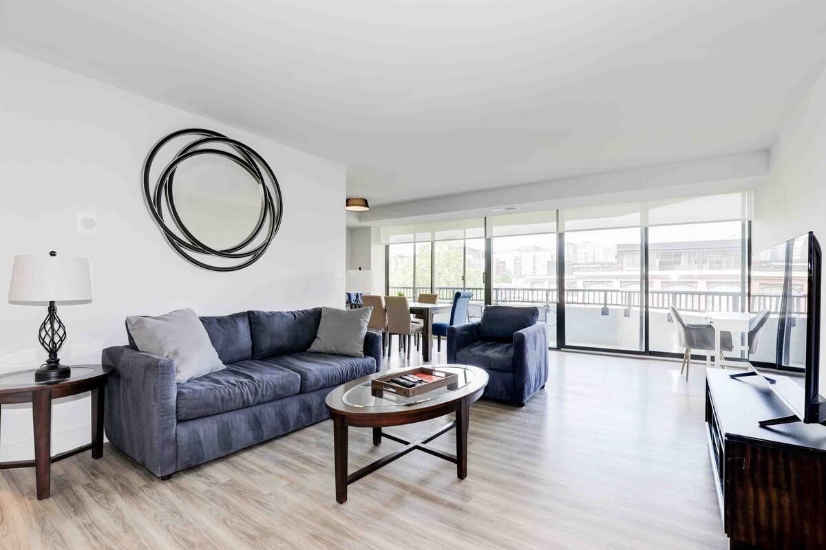 A modern living room with a grey sofa, a coffee table, and a TV stand. There is a large window providing a view of the city.