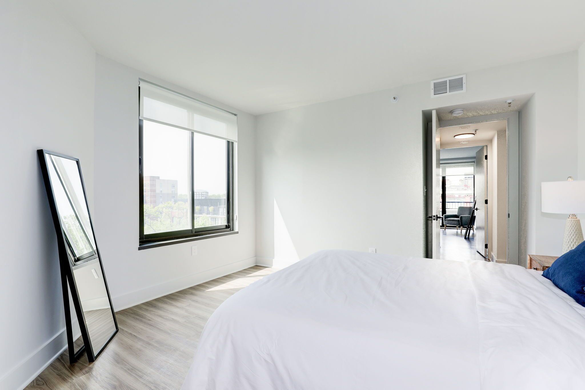 A modern hotel room with a large bed, a window with blinds, a door leading to a hallway, and a view of the city.