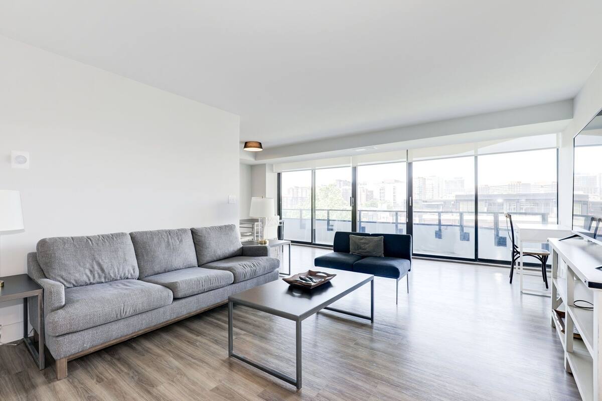 A modern living room with a grey sofa, a coffee table, and a blue sofa. There is a large window providing a view of the cityscape.