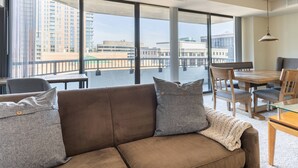 A modern living room with a brown sofa, grey cushions, and a glass coffee table. Large windows offer a view of the cityscape. - Premium A-Location Apartment (Arlington)