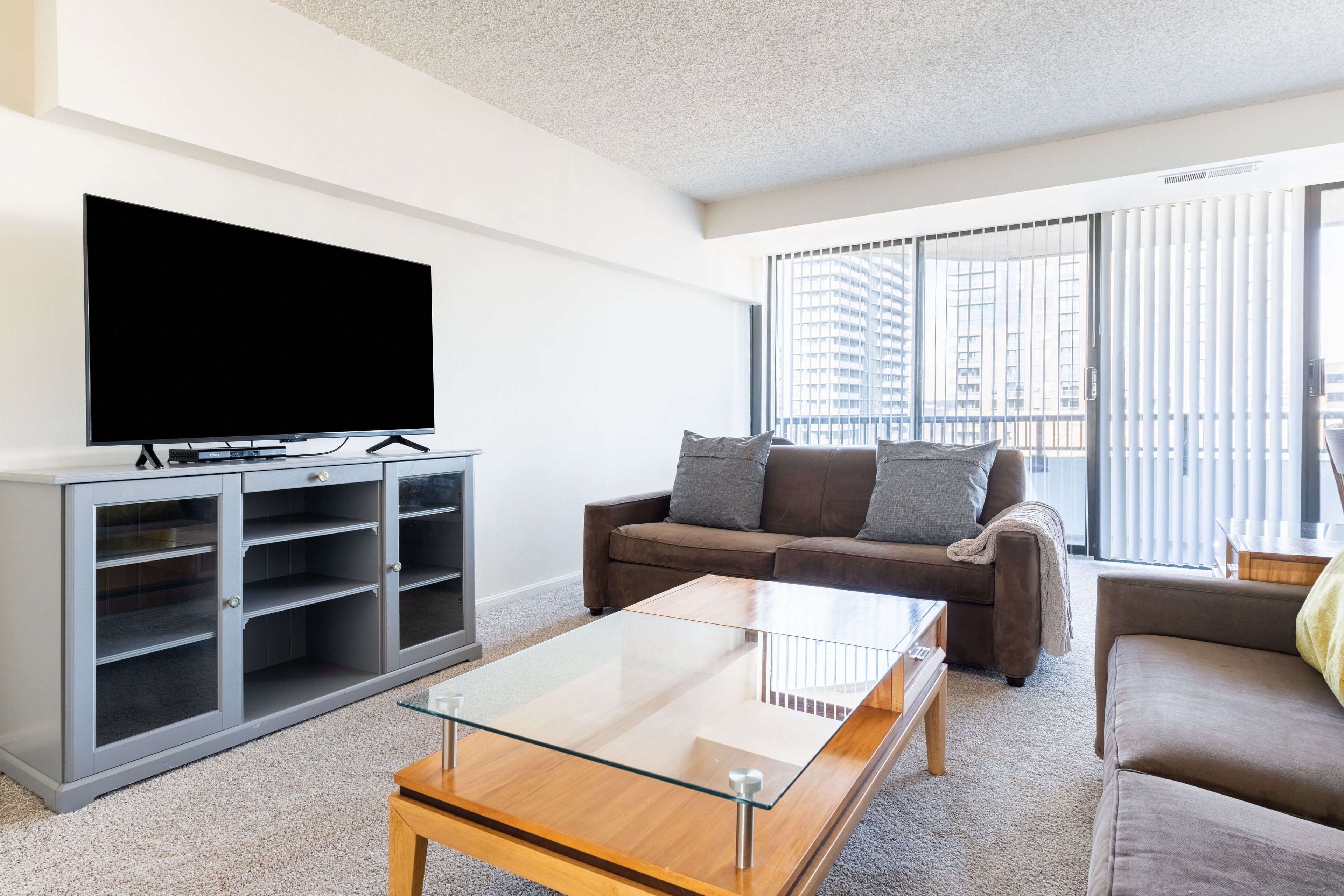 A modern living room with a flat-screen TV, a glass coffee table, a grey entertainment unit, and a brown sofa.