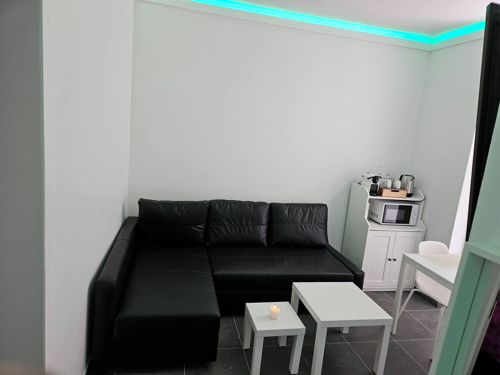 Un salon moderne avec un canapé d’angle noir, une petite table blanche sur laquelle est posée une bougie, et un four à micro-ondes sur un meuble.
