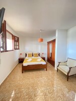 A spacious bedroom with a large bed, a wooden wardrobe, a chair, and a window with blinds.