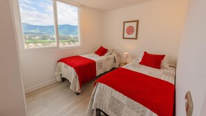 A bedroom with two beds, a window with a view of hills, and a framed picture on the wall. - Oceana Suites Papudo (Papudo)