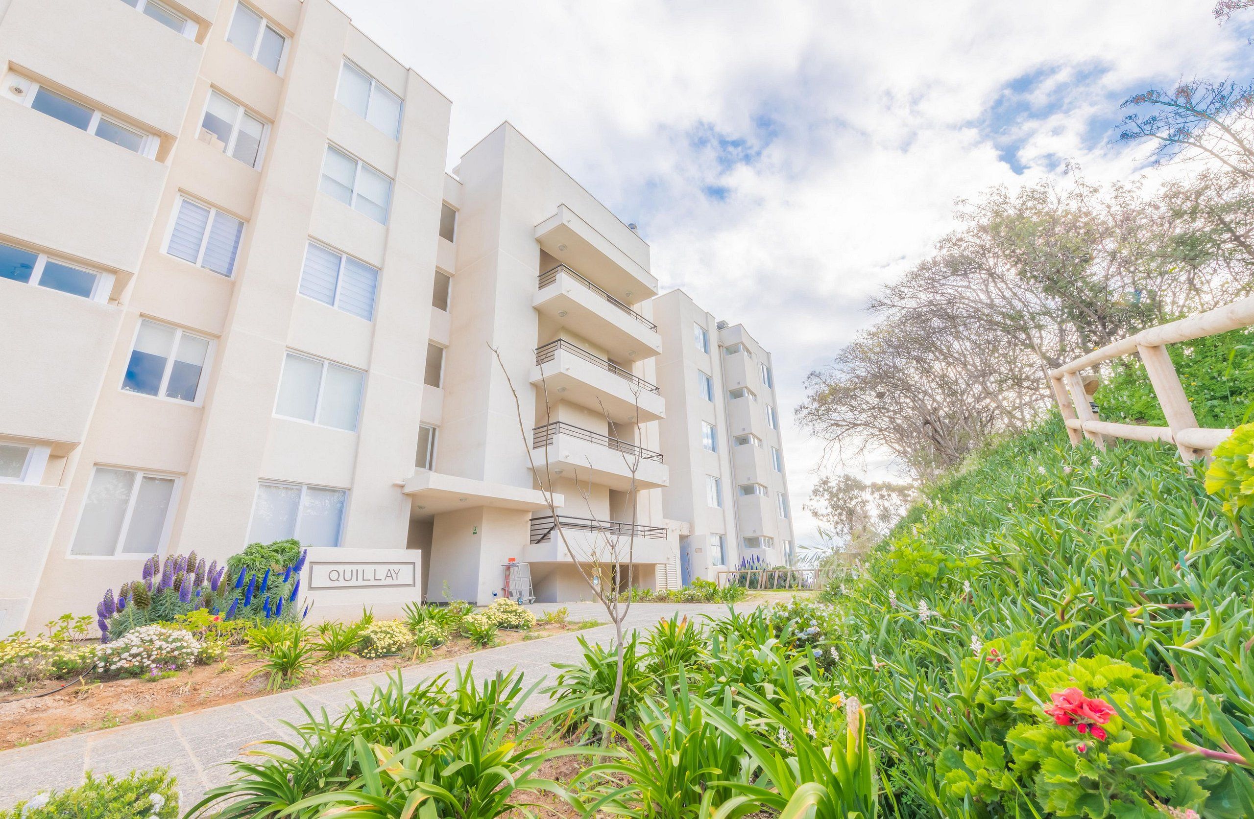 Un edificio residencial de varios pisos con balcones, un cartel que dice 'QUILLAY' y un jardín ajardinado con diversas plantas.