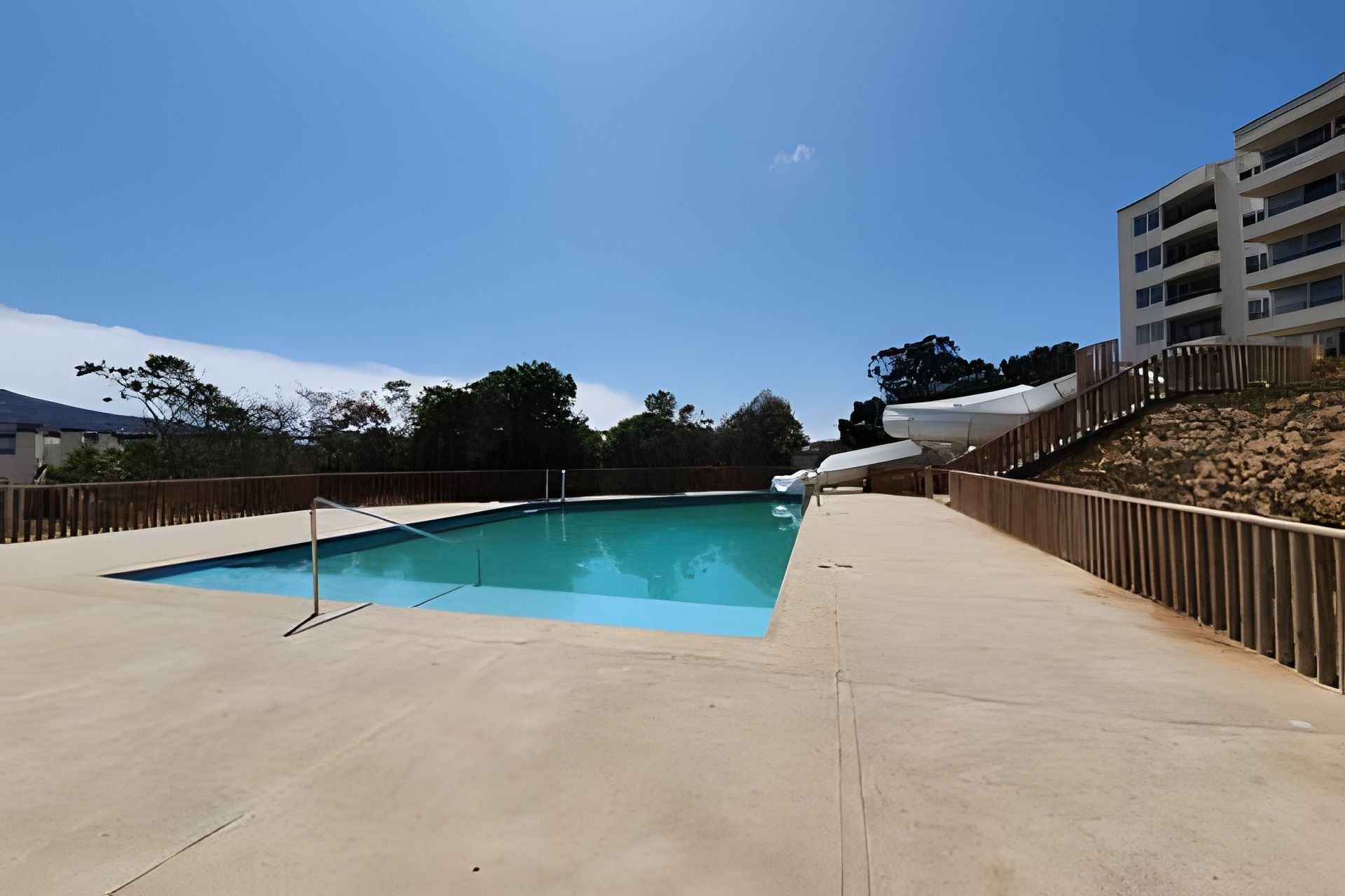 Une piscine avec une terrasse en bois, un parasol blanc et un bâtiment en arrière-plan.