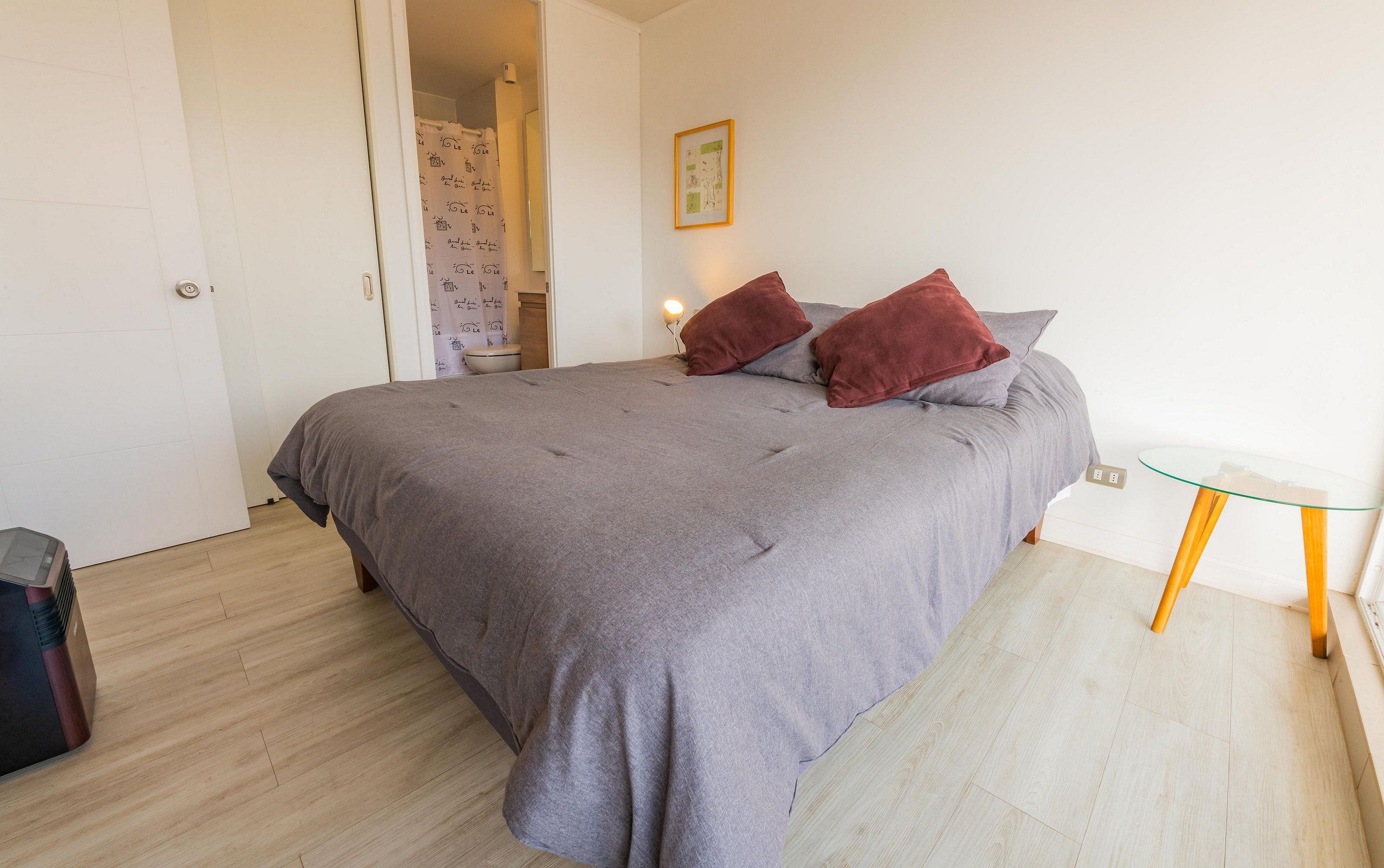 Une chambre à coucher avec un lit, une petite table, une chaise et une salle de bain visible à travers une porte ouverte.