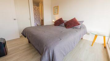 Une chambre à coucher avec un lit, une petite table, une chaise et une salle de bain visible à travers une porte ouverte.