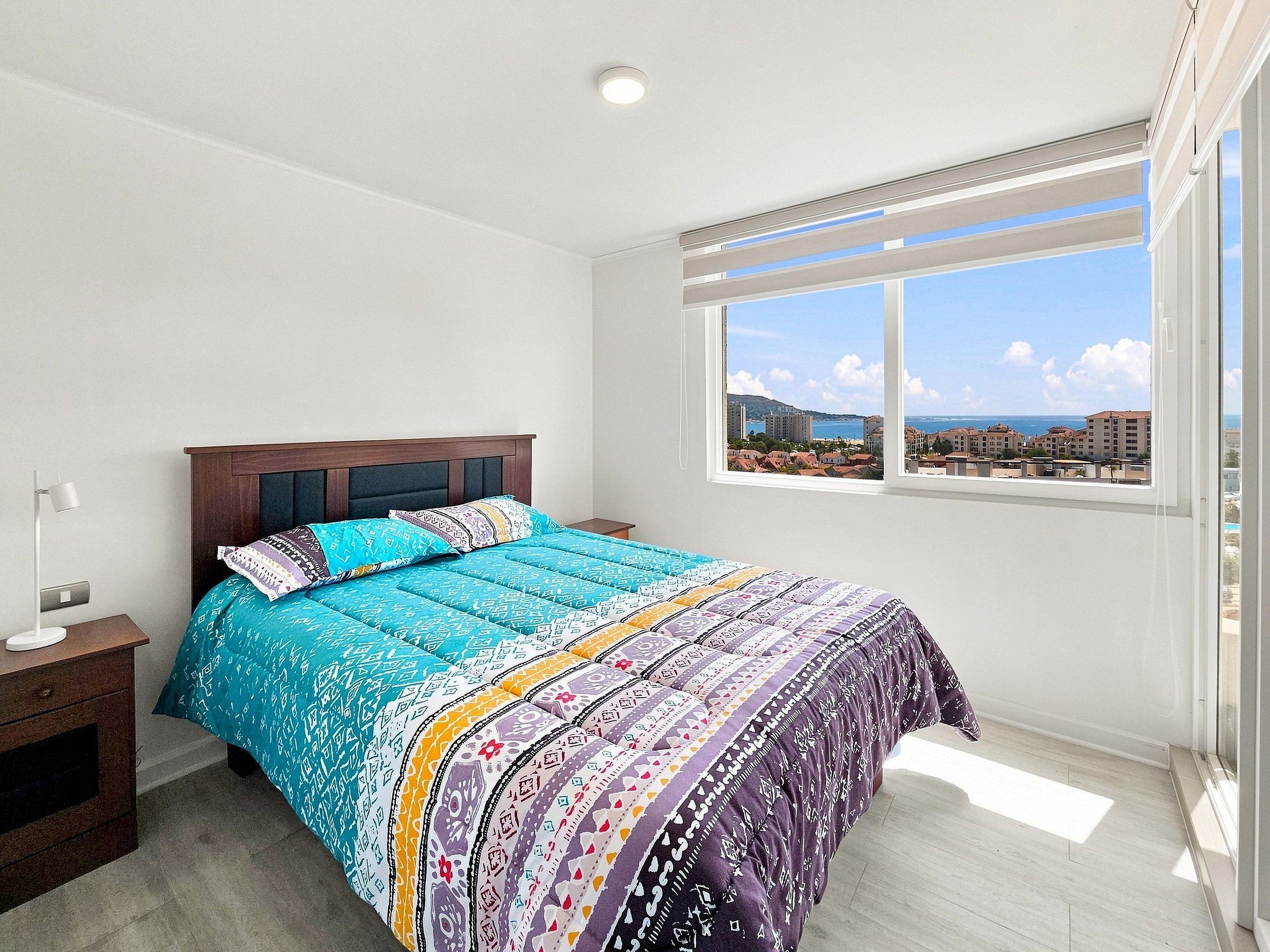A bedroom with a large bed, a nightstand, a window with a view of buildings, and a ceiling light.