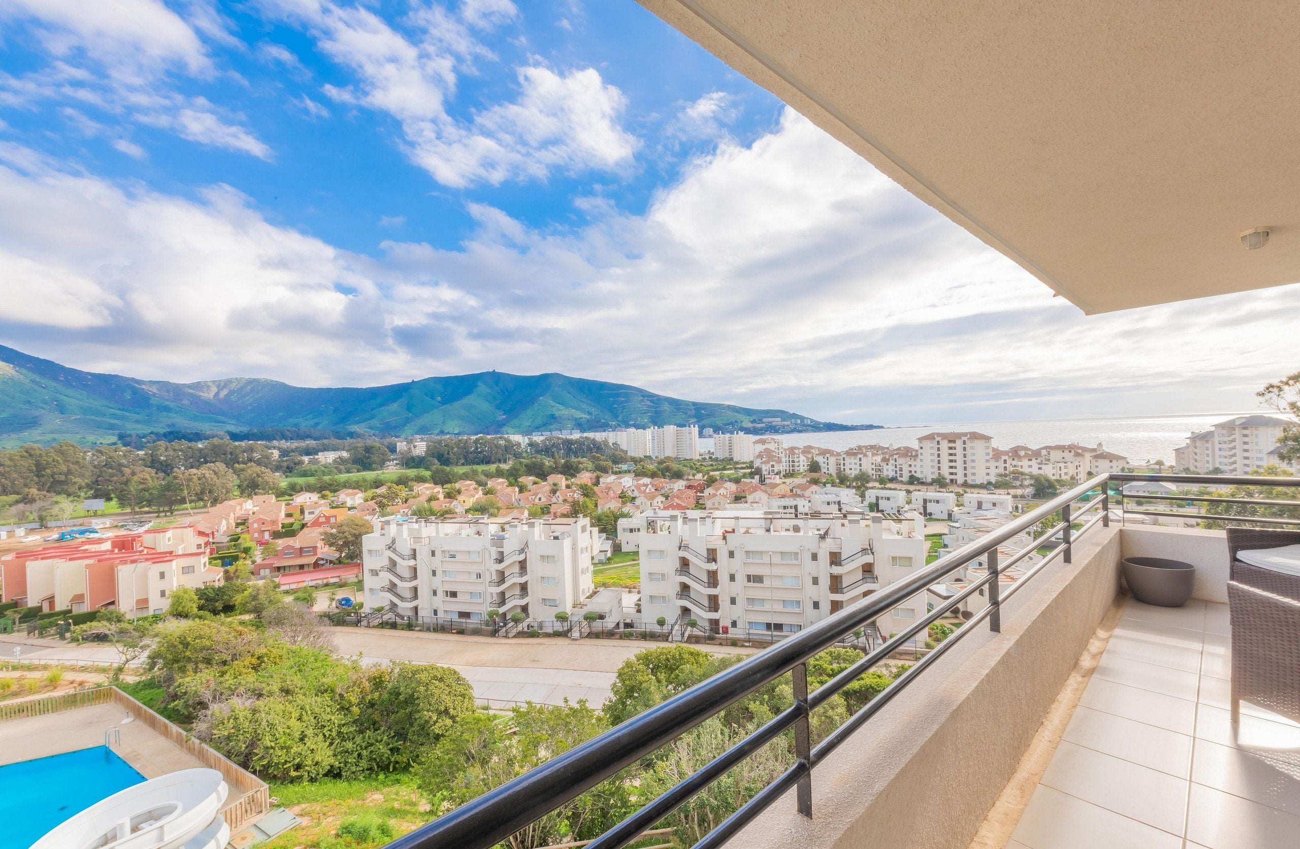 A balcony with a pool, a view of a residential area, and mountains in the distance.