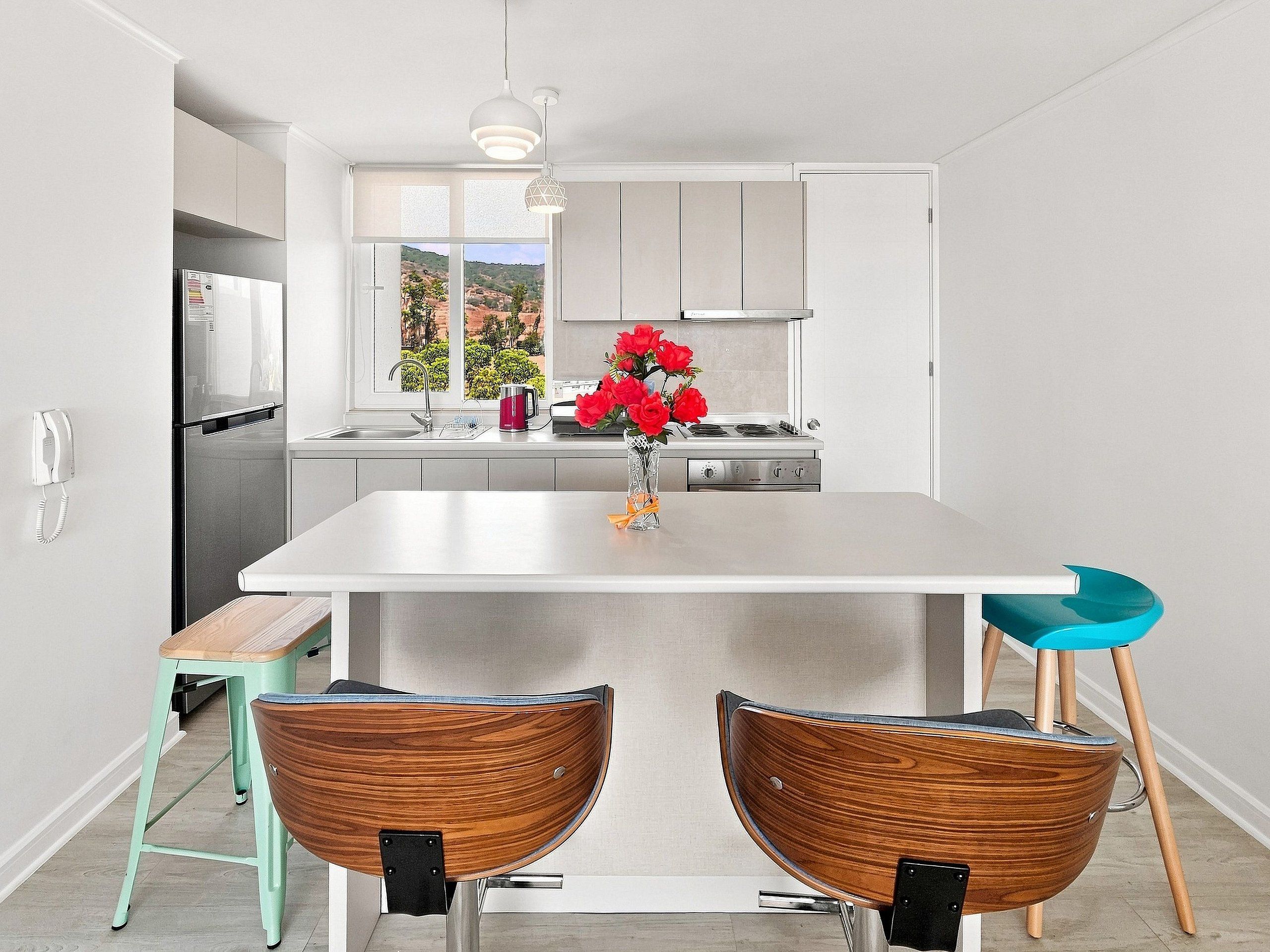 Una cocina moderna con una isla central, dos sillas de madera, un taburete verde azulado y un florero con flores rojas.