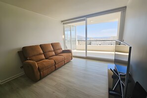 A modern living room with a brown sofa, a glass table, and a large sliding glass door leading to a balcony with a city view.