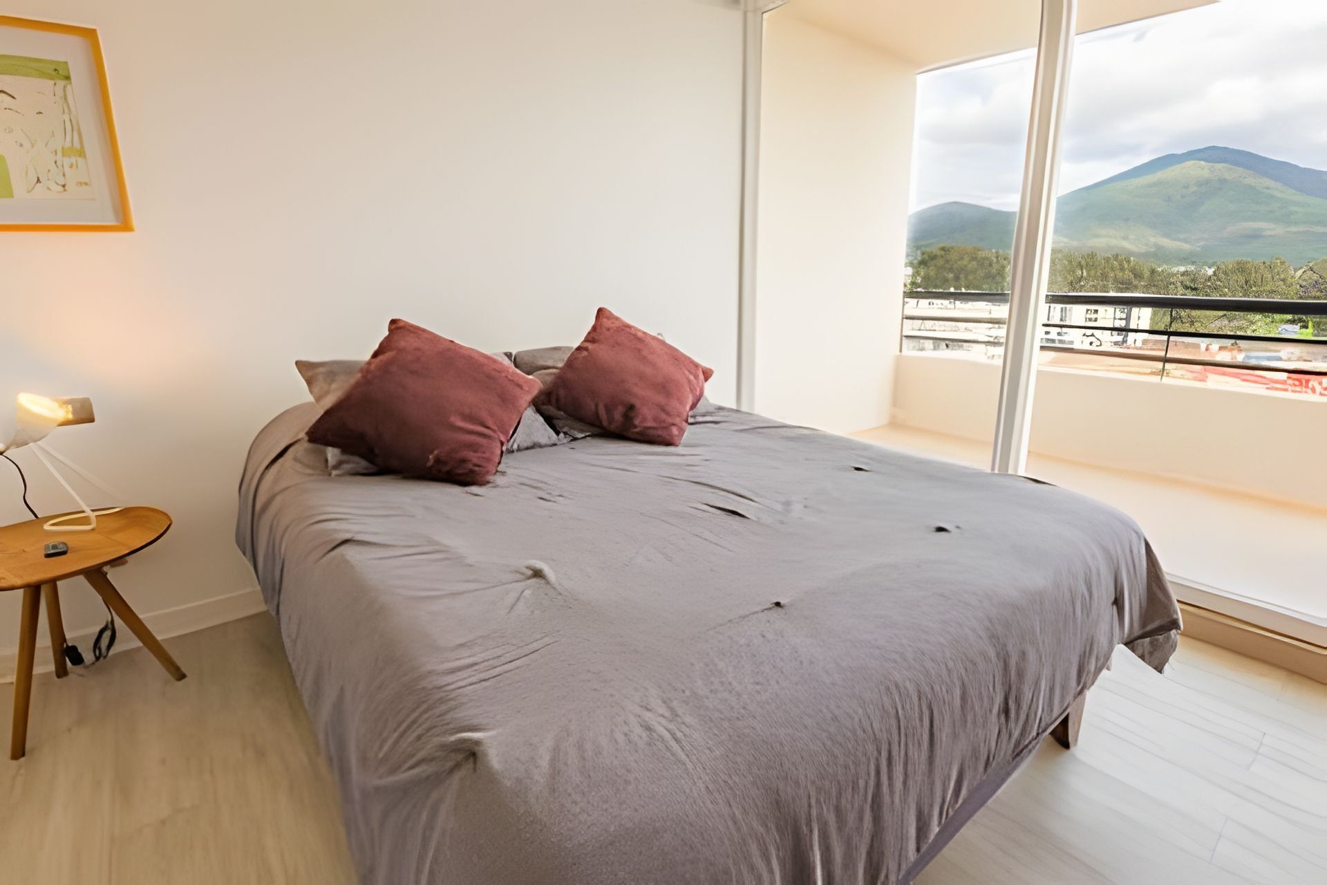A bedroom with a large bed, two pillows, a bedside table with a lamp, and a view of mountains through large windows.