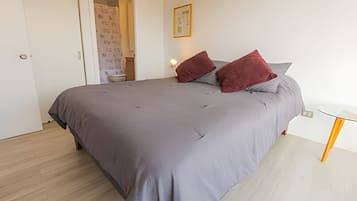 Un dormitori amb un llit gran, una tauleta de nit, una porta que dona a un bany i un quadre al mur.