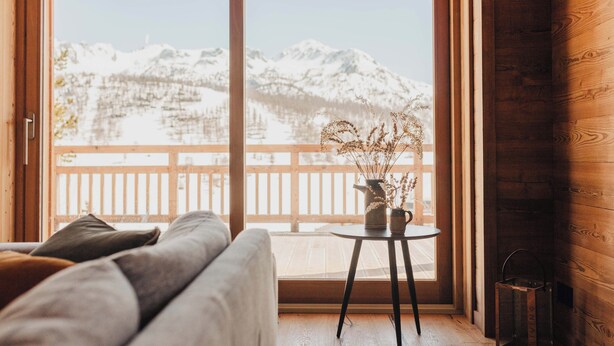 A cozy room with a wooden floor, a sofa, a small table with a vase of dried plants, and a view of snow-covered mountains through large windows. - Chalet La Belle Etoile (Montgenèvre)