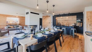 A modern kitchen with wooden cabinets, a dining area with a table set for six, and a living room with a sofa and a large window. - Chalet La Belle Etoile (Montgenèvre)