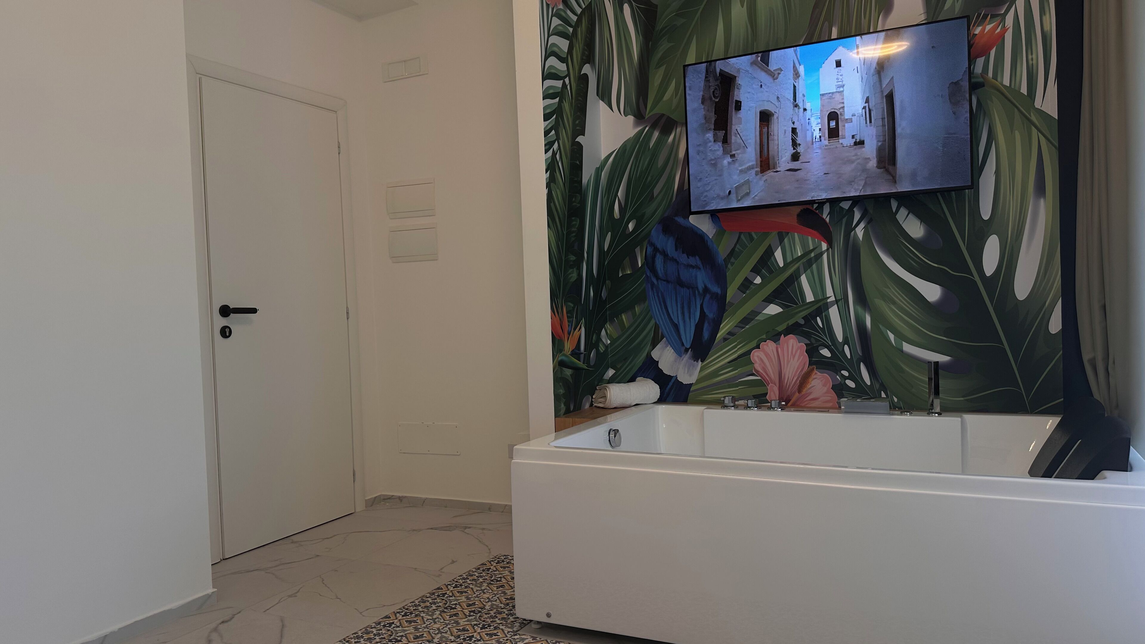 A bathroom with a white bathtub, a television mounted on the wall, and a tropical leaf-patterned mural.