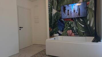 A bathroom with a white bathtub, a television mounted on the wall, and a tropical leaf-patterned mural.