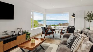 A modern living room with a large window, a flat-screen TV, and a wooden coffee table. - Pen Gar - Elevated Sea Views, EV Charger (Saundersfoot)