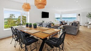 A spacious living room with a large wooden dining table, black chairs, and a grey sofa. There are two large windows offering a view of the outdoors. - Pen Gar - Elevated Sea Views, EV Charger (Saundersfoot)