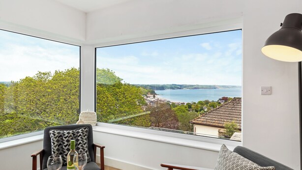 A room with a large window offering a view of a lake and greenery. There is a table with a bottle and glasses, and a lamp on the wall. - Pen Gar - Elevated Sea Views, EV Charger (Saundersfoot)