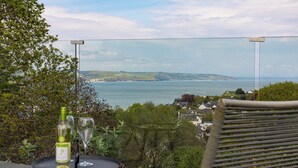 A table with a bottle and a glass, overlooking a coastal landscape with houses and greenery. - Pen Gar - Elevated Sea Views, EV Charger (Saundersfoot)