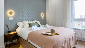 A bedroom with a bed, bedside tables, a window, and a view of buildings outside.