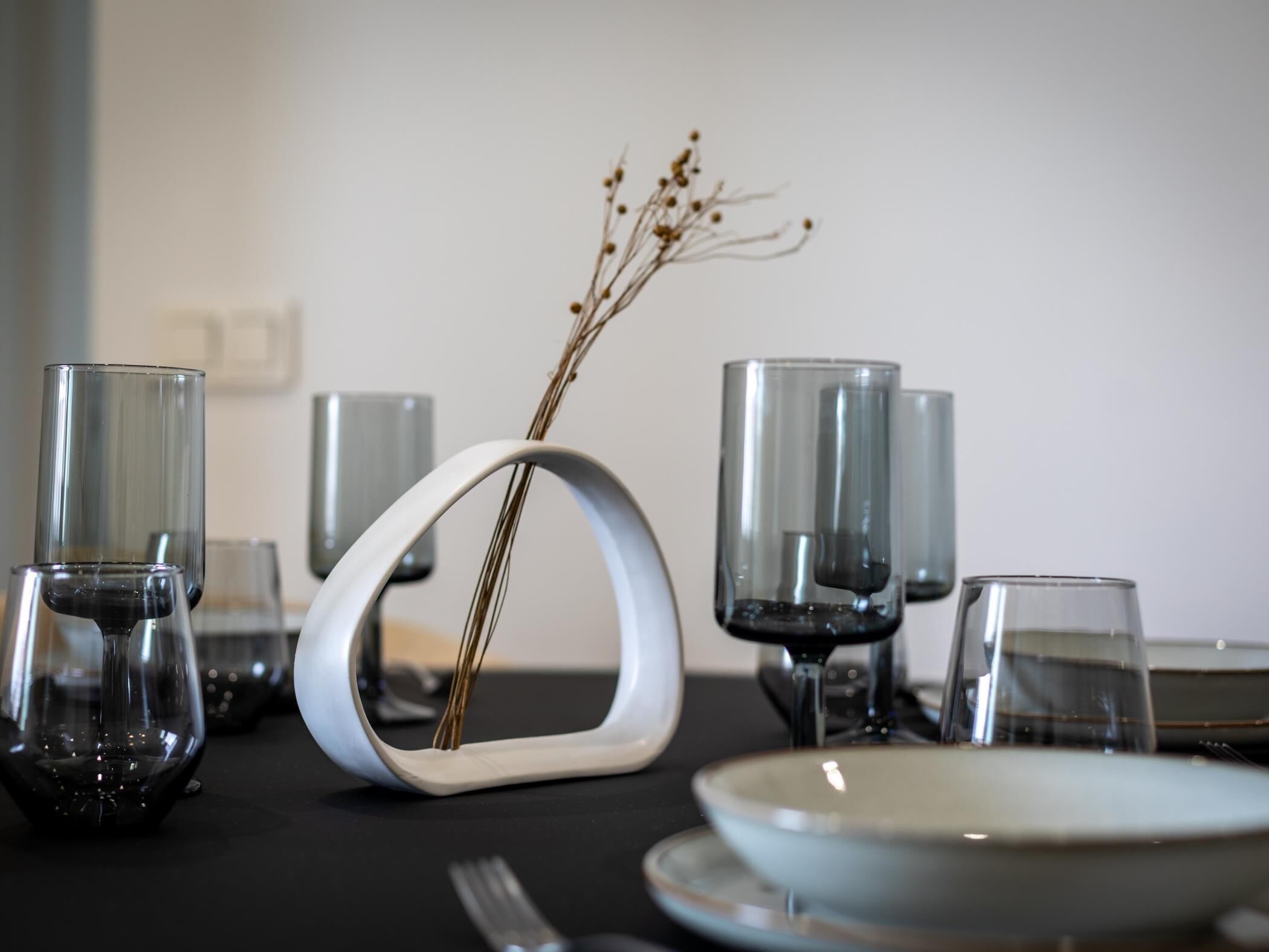 A dining table set with clear glassware, a white vase with dried plants, and a plate.