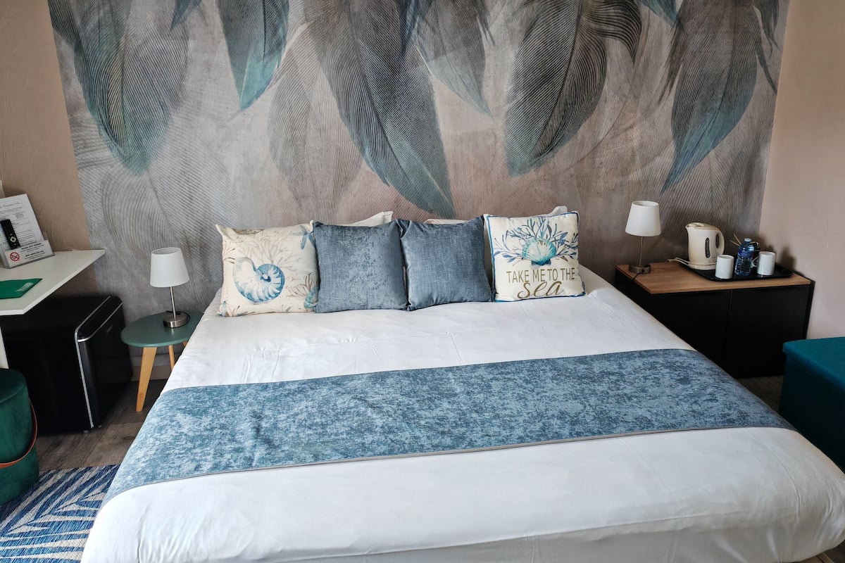 A hotel room with a large bed, a desk, a chair, a nightstand, a lamp, a coffee maker, and a water bottle.