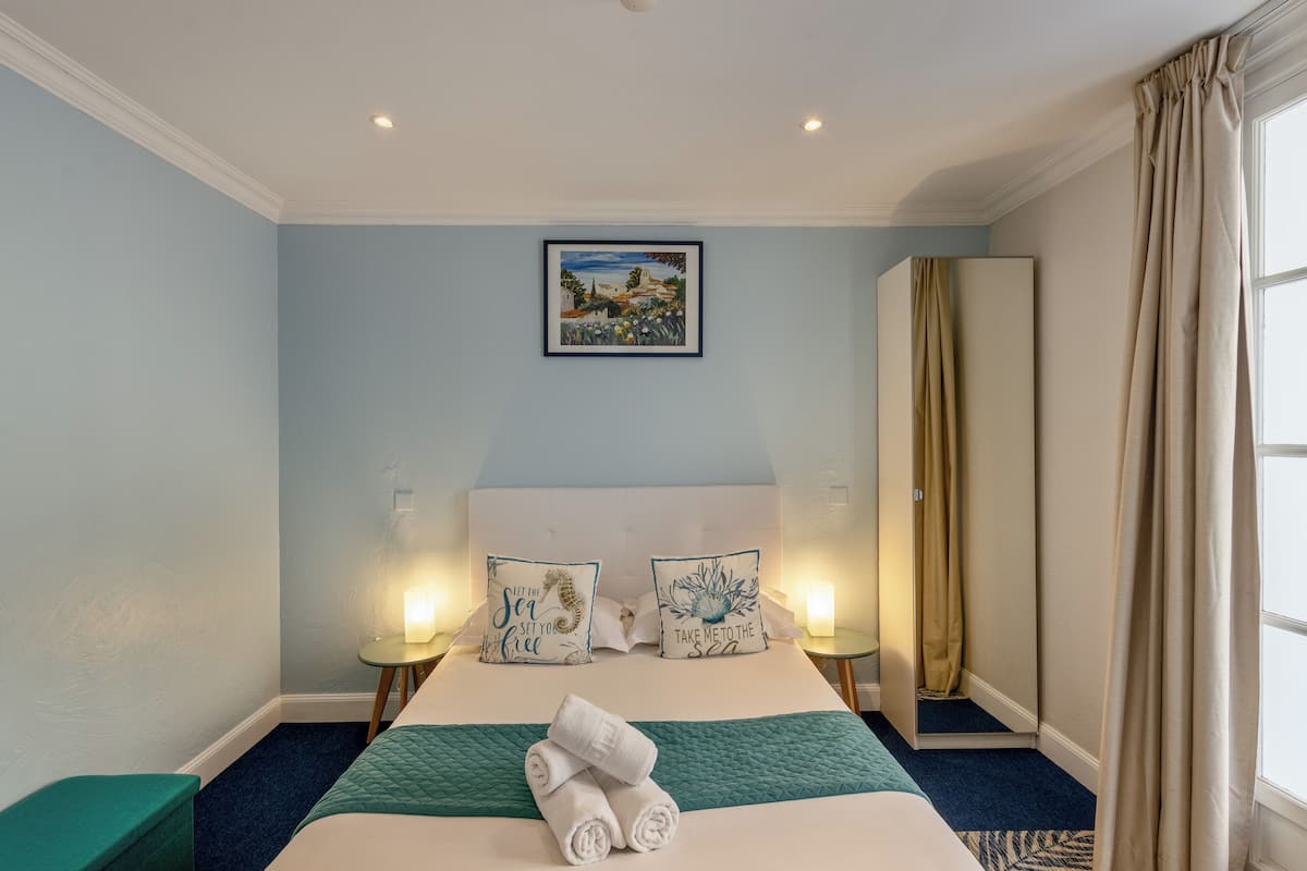 A hotel room with a bed, two bedside tables with lamps, a painting on the wall, and a view of the sea through a window.