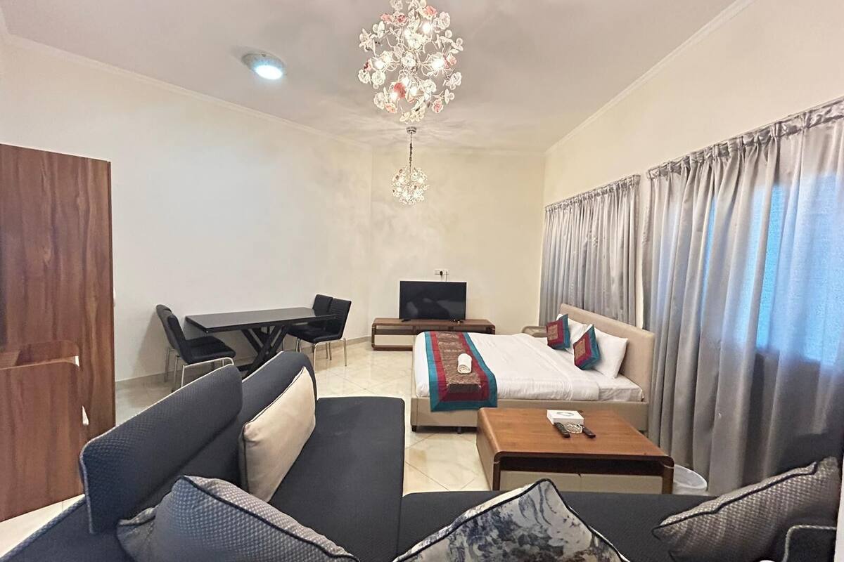A hotel room with a sofa set, a bed with a blanket and pillows, a TV, a desk with a chair, and a chandelier.