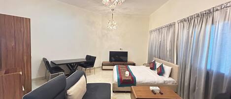 A hotel room with a sofa set, a bed with a blanket and pillows, a TV, a desk with a chair, and a chandelier.
