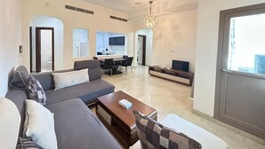 Una sala de estar moderna con sofá, mesa de centro y televisor. Se observa un área de cocina a través de un separador de vidrio.