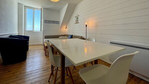 A modern dining area with a white table, white chairs, a black sofa, and a hanging light fixture. - "les Yeux en Bleu"- 2 ch- Magnifique vue sur la Grande Plage (Quiberon)