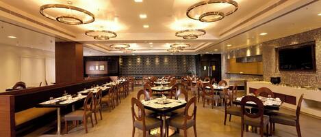 A spacious restaurant with multiple dining tables and booths, modern lighting fixtures, and a large flat-screen TV on the wall.