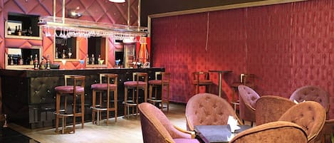 Eine Bar mit roter Wand, Muster, mehreren Barhocker und einer Bar, die mit Flaschen und Gläsern bestückt ist.