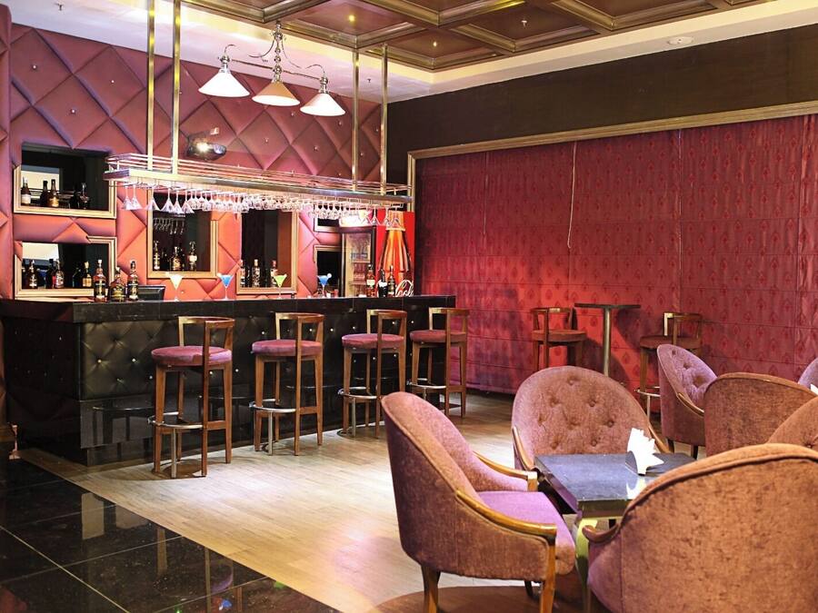 A bar area with a red patterned wall, multiple bar stools, and a bar counter with bottles and glasses.
