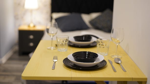 A neatly set dining table with plates, cutlery, and glasses, positioned in a room with a bed and nightstand in the background.