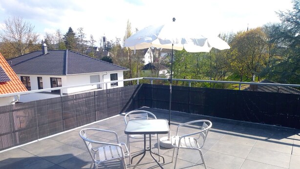 Terraza en la azotea con una mesa, sillas, un paraguas blanco y vistas a casas y árboles.