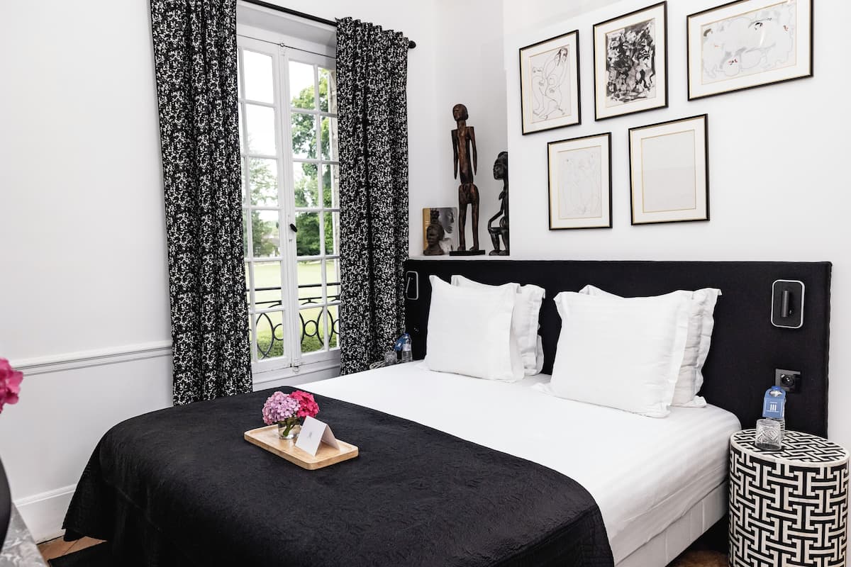 Quarto com colcha preta e branca, mesa de cabeceira de madeira com um copo e uma janela com cortinas florais em preto e branco.
