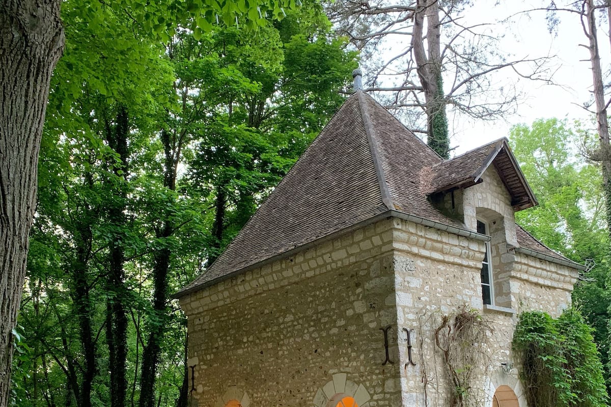 Edifício de pedra com telhado de telhas, rodeado por árvores. Há mesas e cadeiras ao ar livre, e um cão é visível.